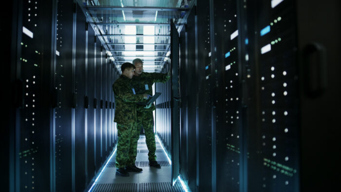 In Data Center Two Military Men Work With Open Server Rack Cabinet. One Holds Military Edition Laptop.